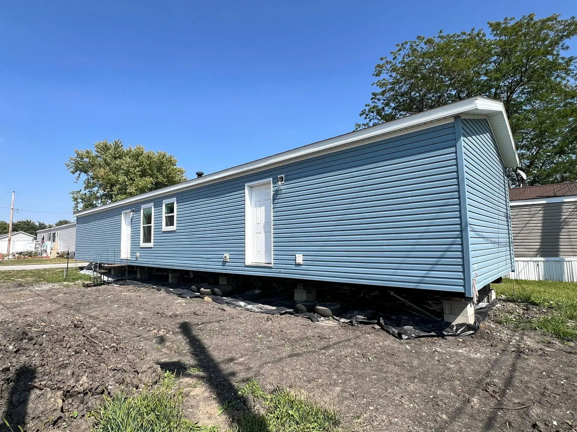 Blue mobile home with white trim on a dirt lot under a sunny blue sky.