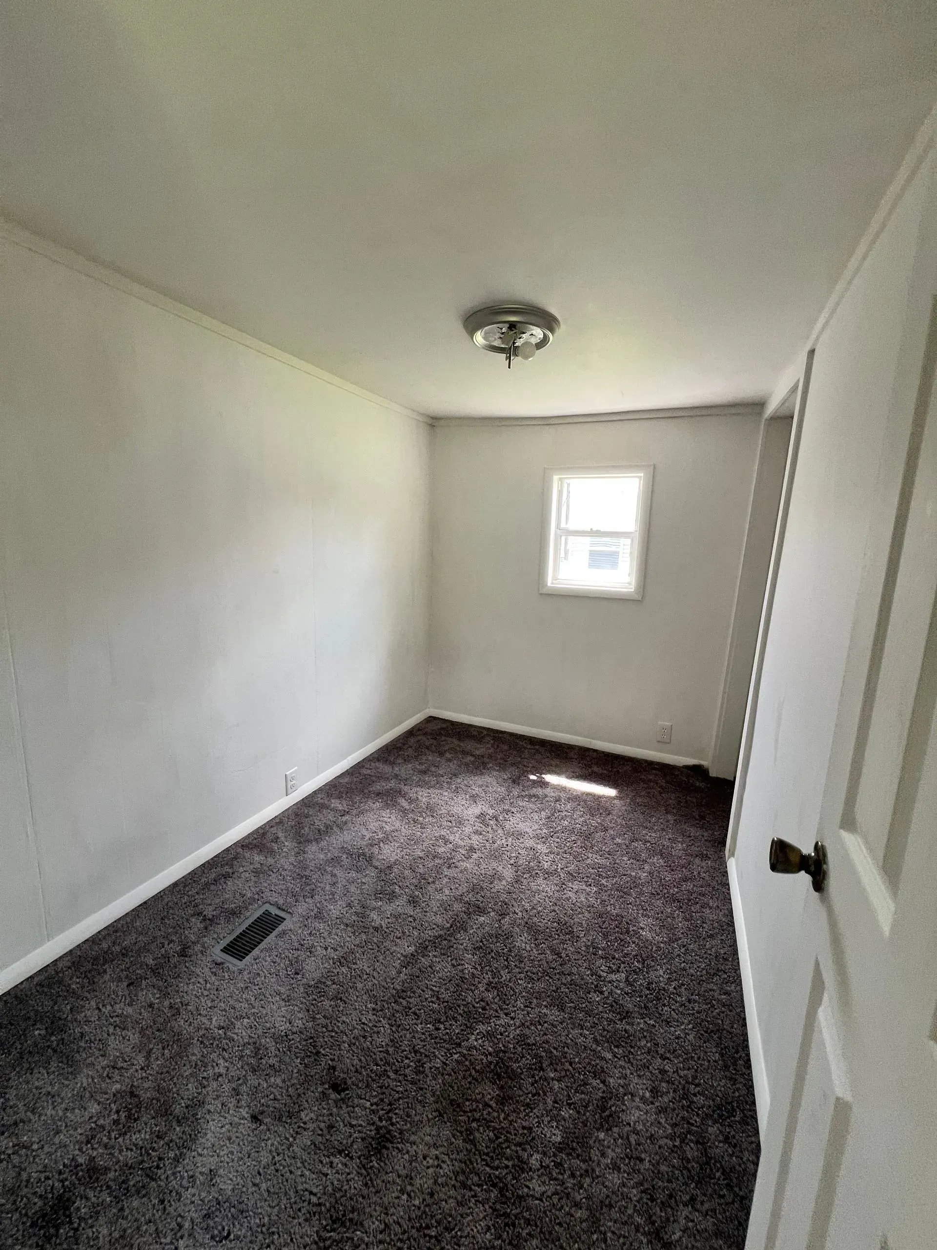 Empty bedroom with dark brown carpet, white walls, and small window.