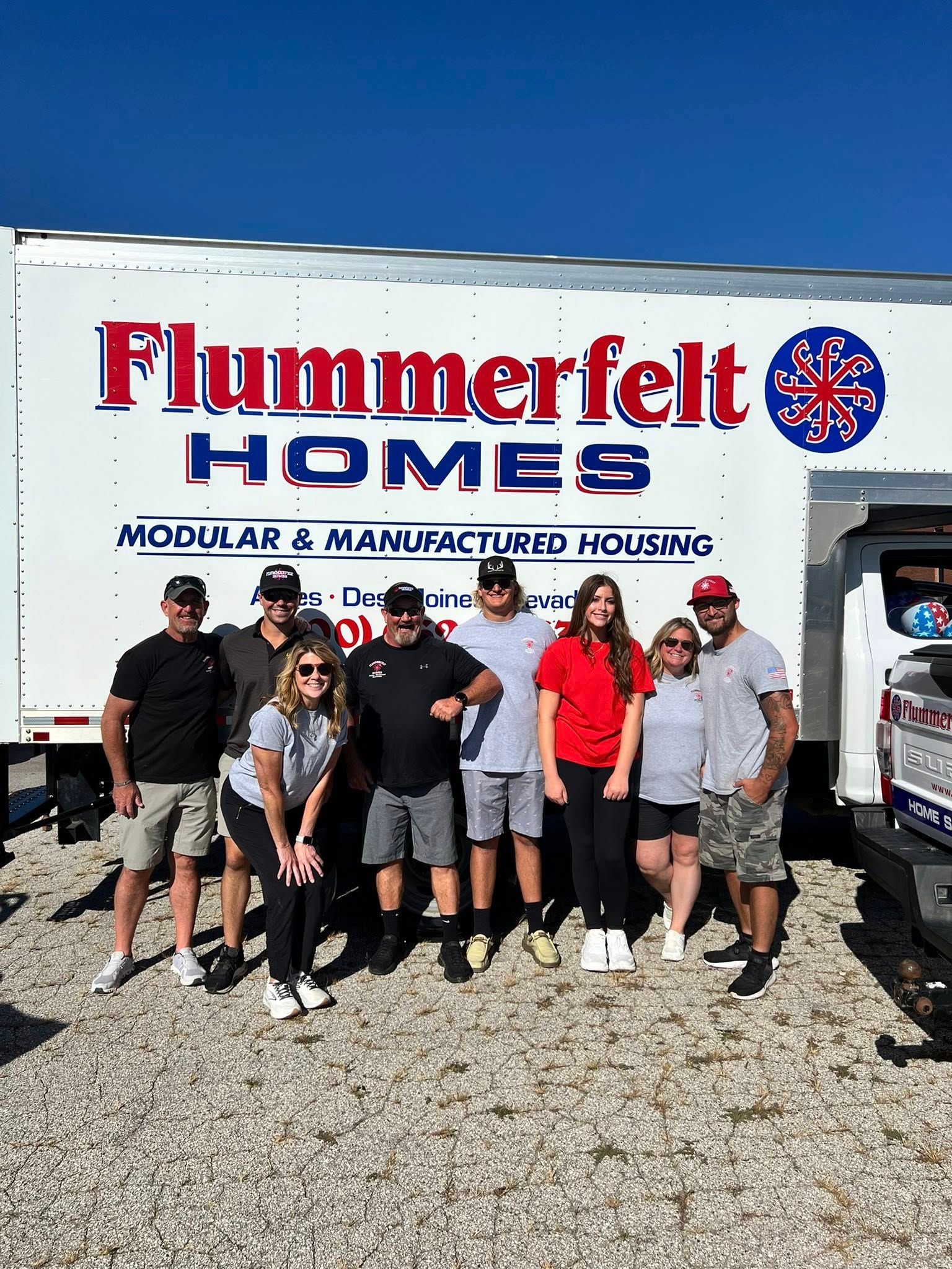 Group of people standing in front of a white truck with "Flummerfelt Homes" logo. Blue sky background.