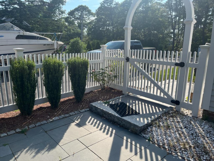 White picket fence with a gate and three tall, green bushes. Gray stone pathway.