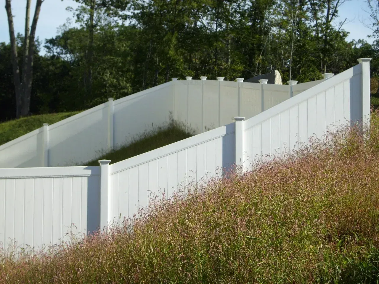 A white fence surrounds a grassy hillside with trees in the background