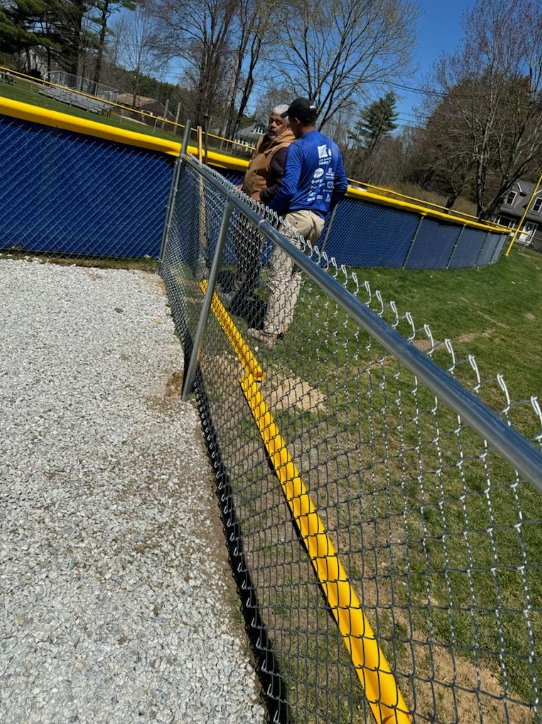 Two men by a chain link fence bordering a baseball field with blue padded wall and yellow trim.