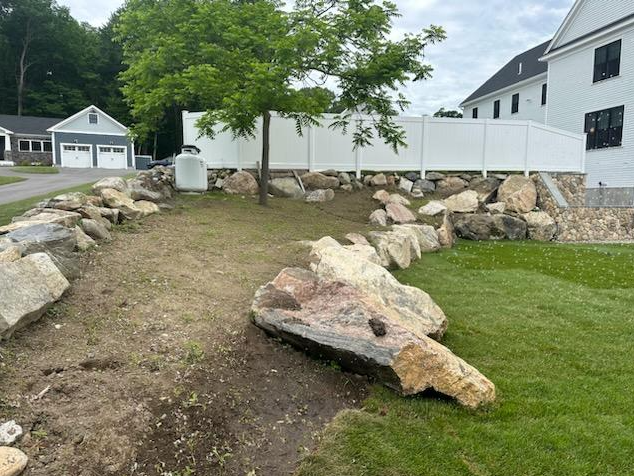 Large rocks border a grassy yard; white fence and two-story building in the background.