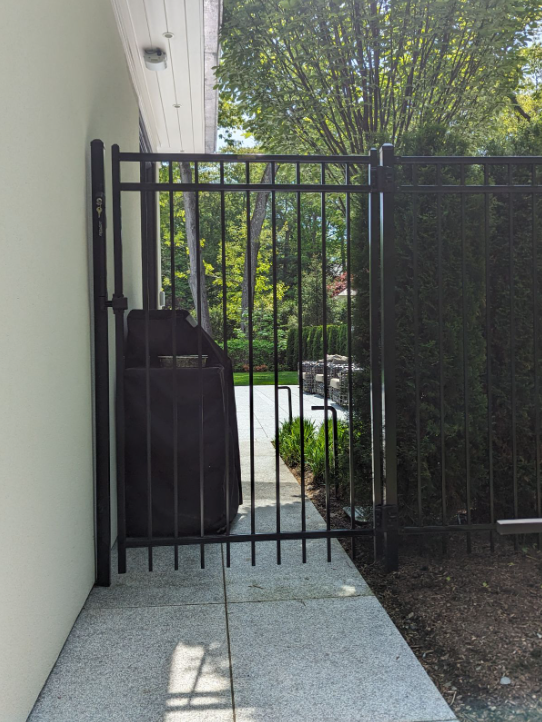 Black metal gate in a paved area, next to a white wall, leading to a yard with greenery.