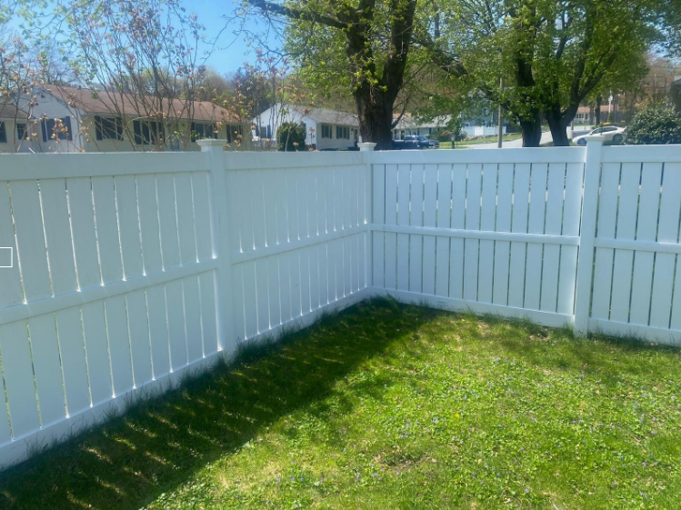 White picket fence corner surrounding green grass lawn, with houses and trees in the background.