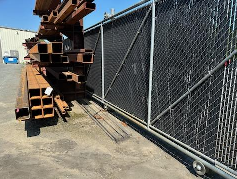Rusty steel beams stacked outdoors next to a chain-link fence.