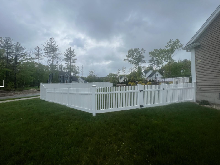 White picket fence enclosing a grassy yard next to a house, under a cloudy sky.