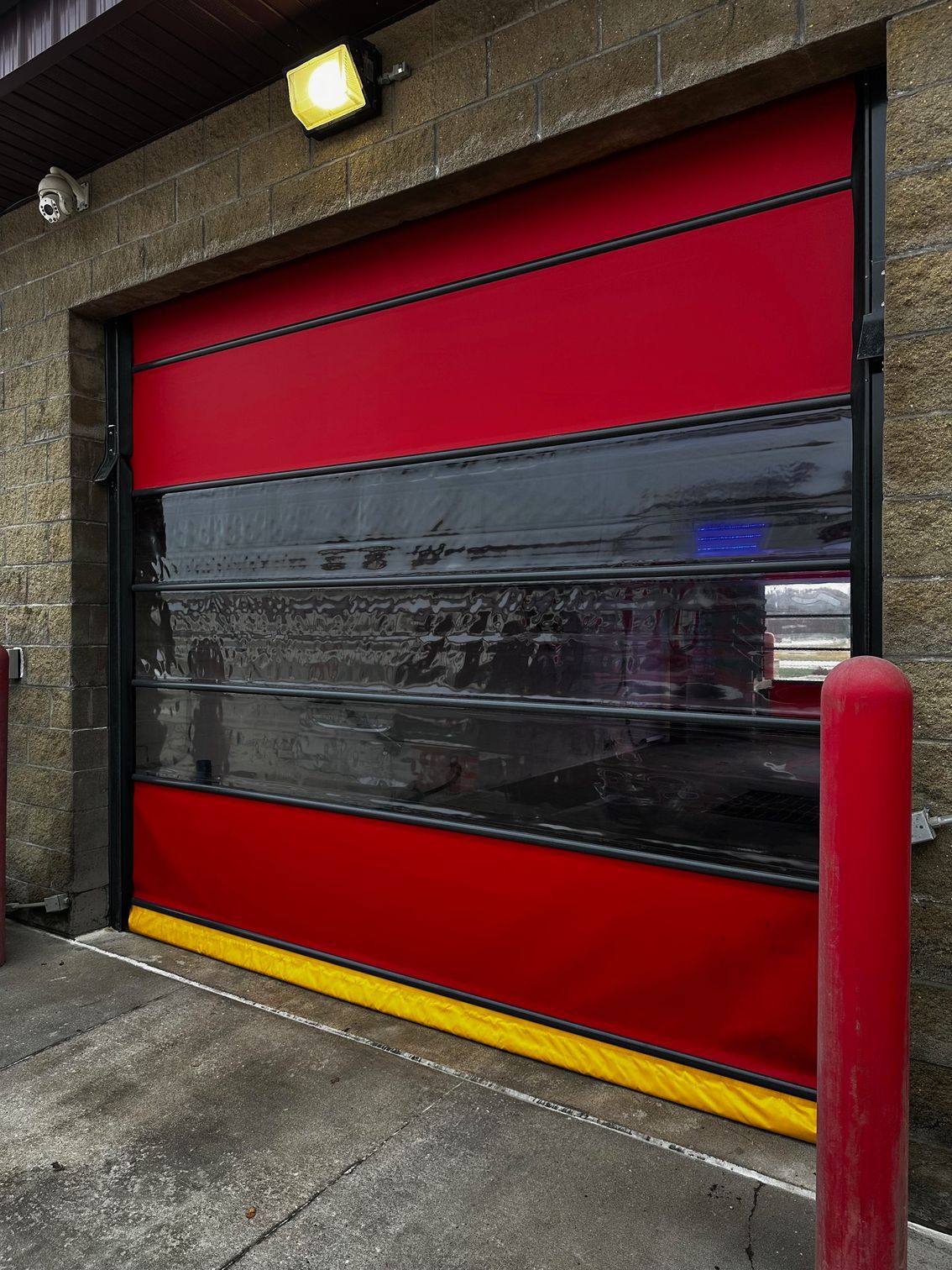Red and black industrial roll-up door with yellow bumper, mounted on a brick building with a light fixture.