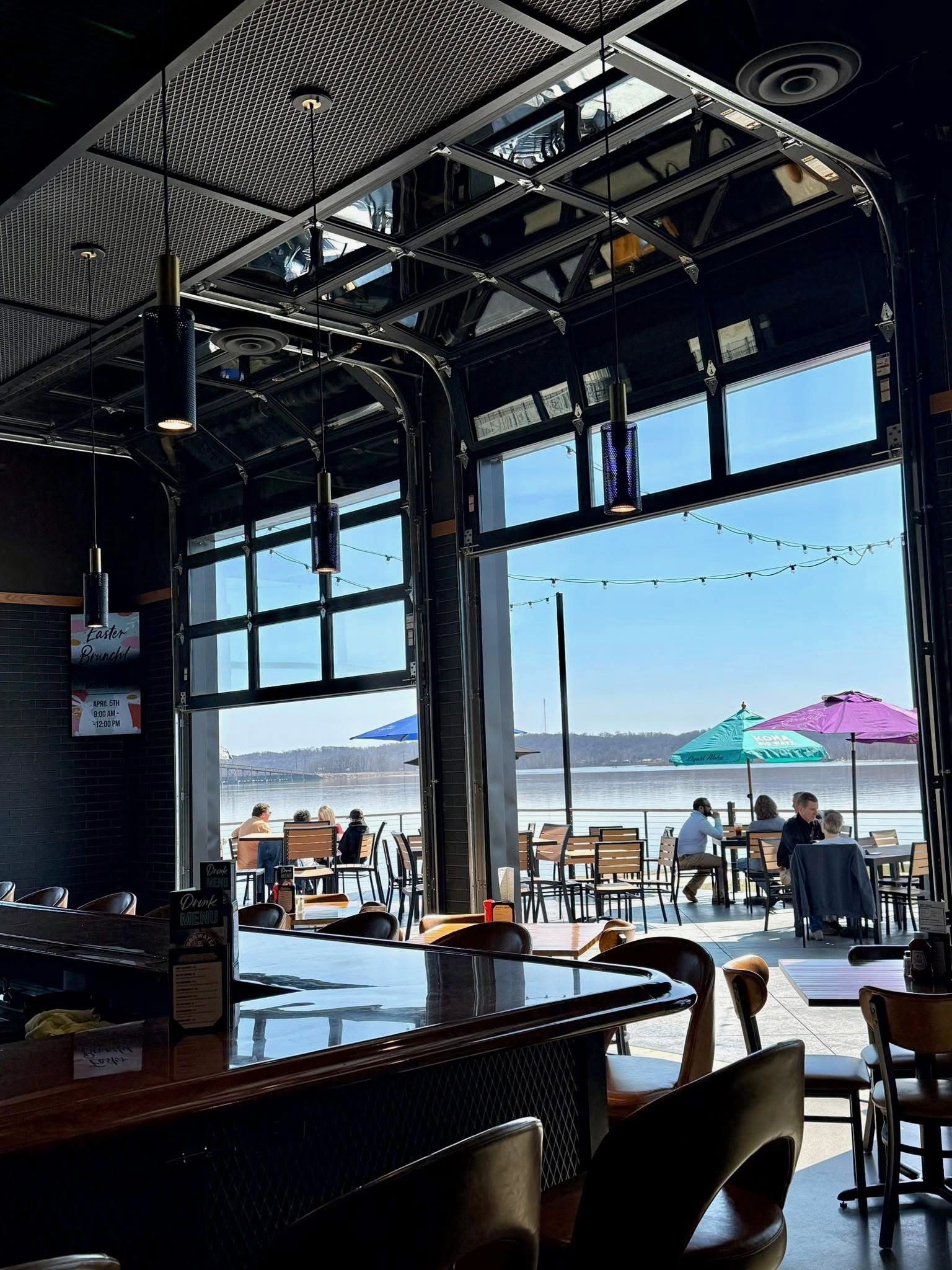 A view from inside a bar looking through an open garage-style door at a patio with colorful umbrellas overlooking water.