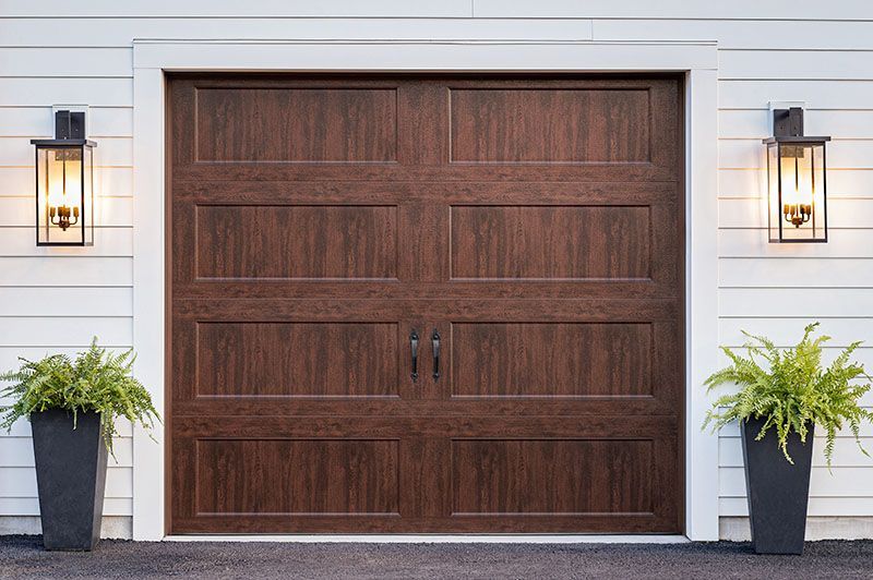 Brown garage door with dark accents, flanked by ferns in black planters and wall-mounted lights on a white building