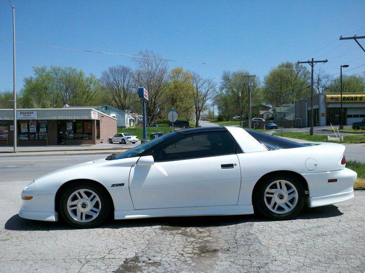White sports coupe parked on a street under a clear sky, side view with storefronts in the background