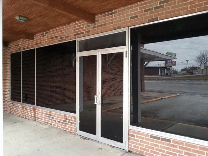 Storefront with dark-tinted windows and a glass door under a brick overhang, seen from outside.