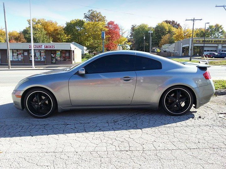 Silver sports coupe parked on a cracked lot with tinted windows and black rims, buildings and autumn trees behind it