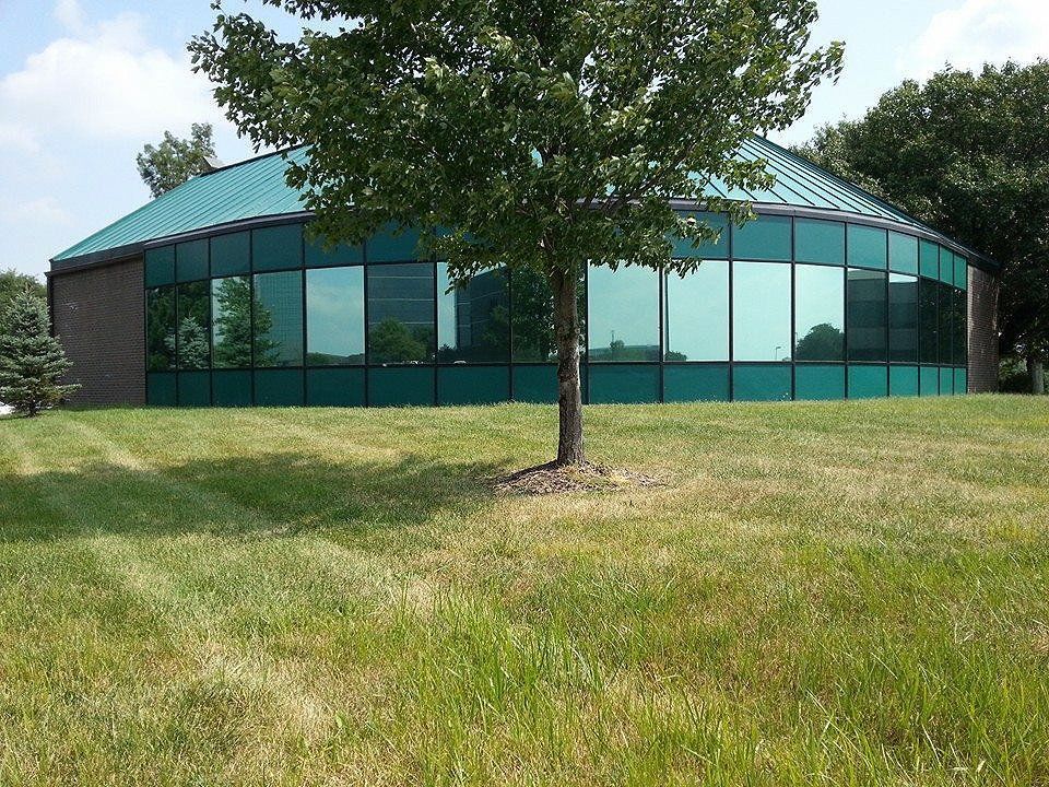 Low, round building with teal roof and tinted windows in a grassy field, with a tree in front.