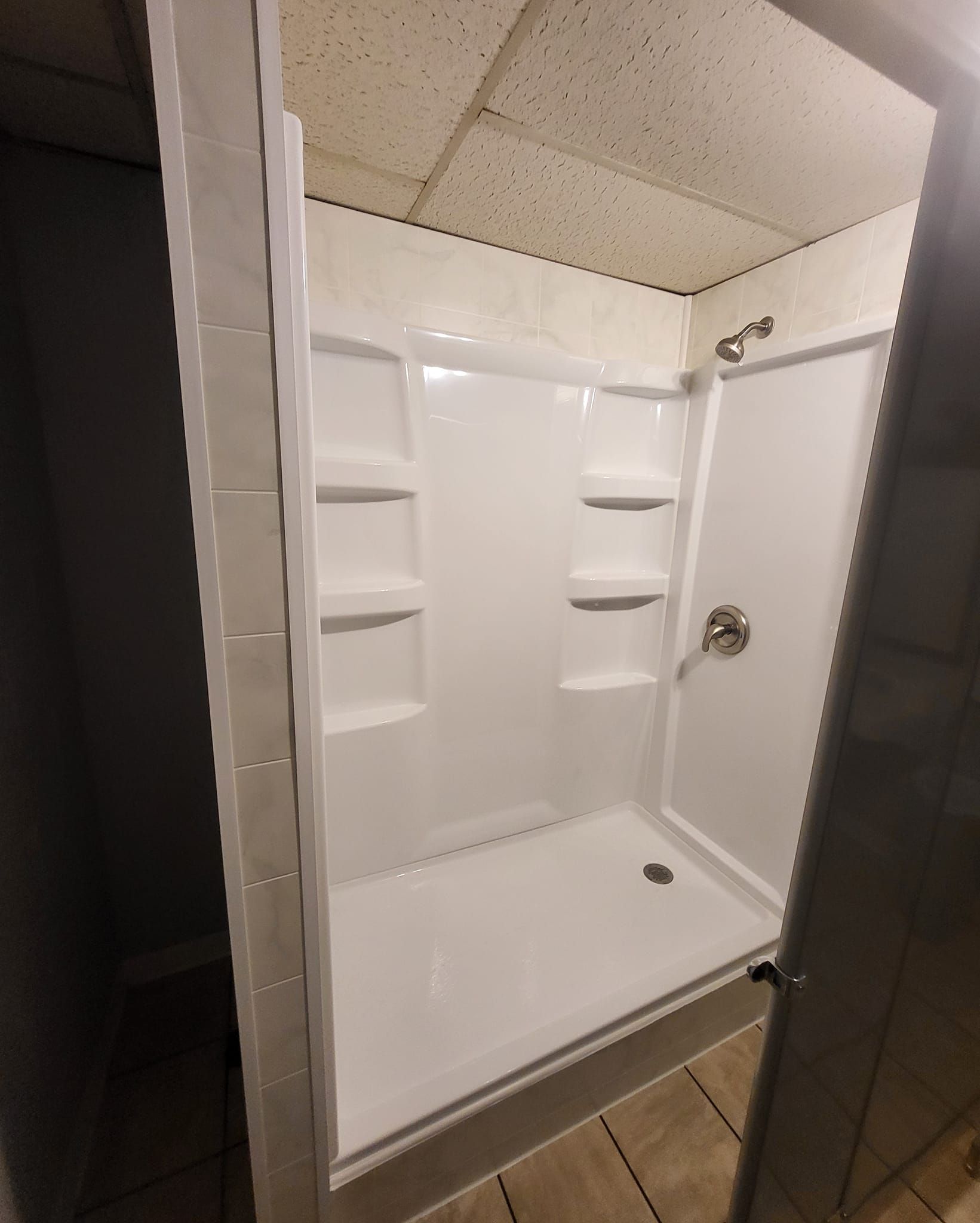 White shower stall with shelves, chrome fixtures, and beige tile in a bathroom.