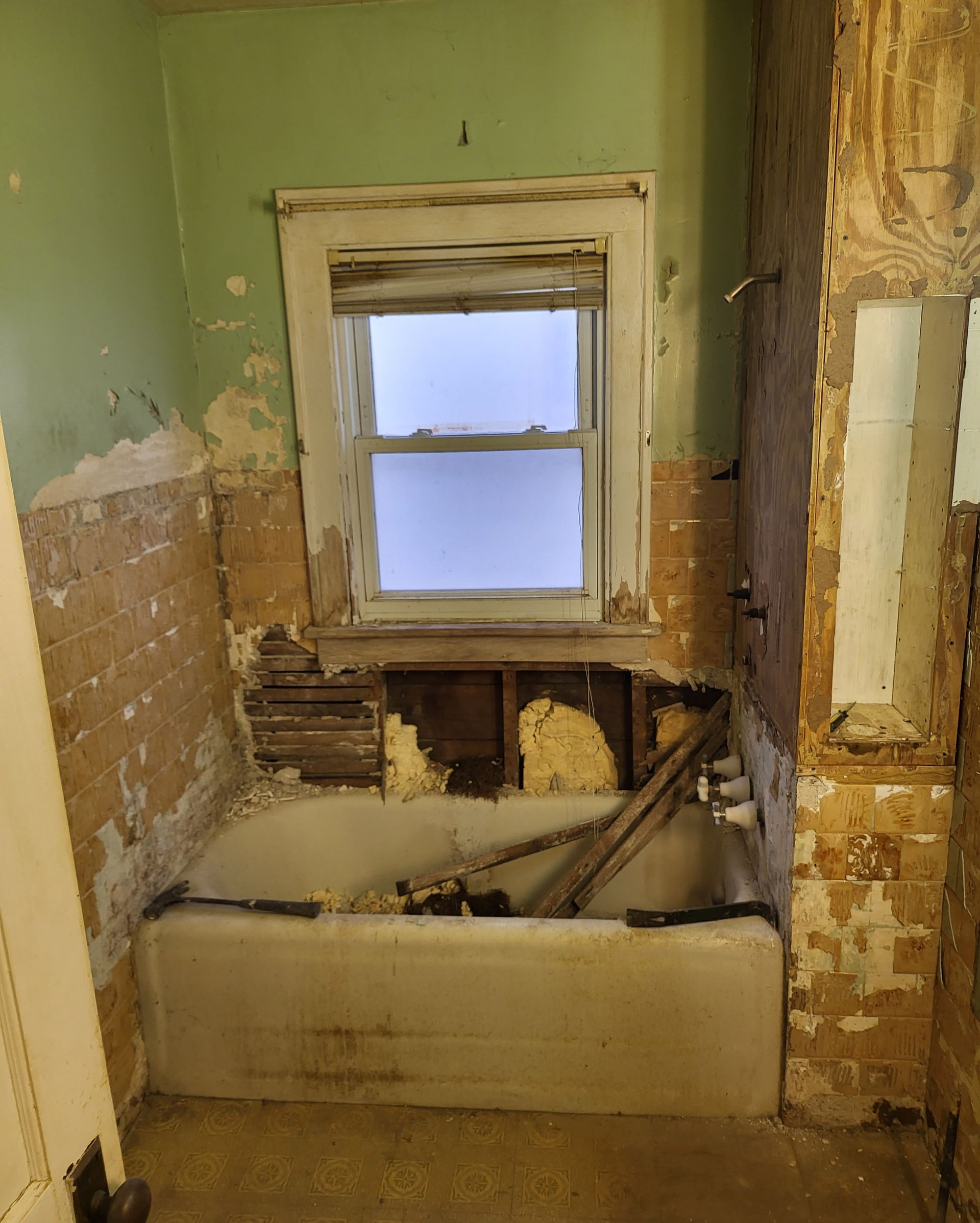 Bathroom undergoing renovation, showing a tub under a window; walls stripped of tile and plaster.