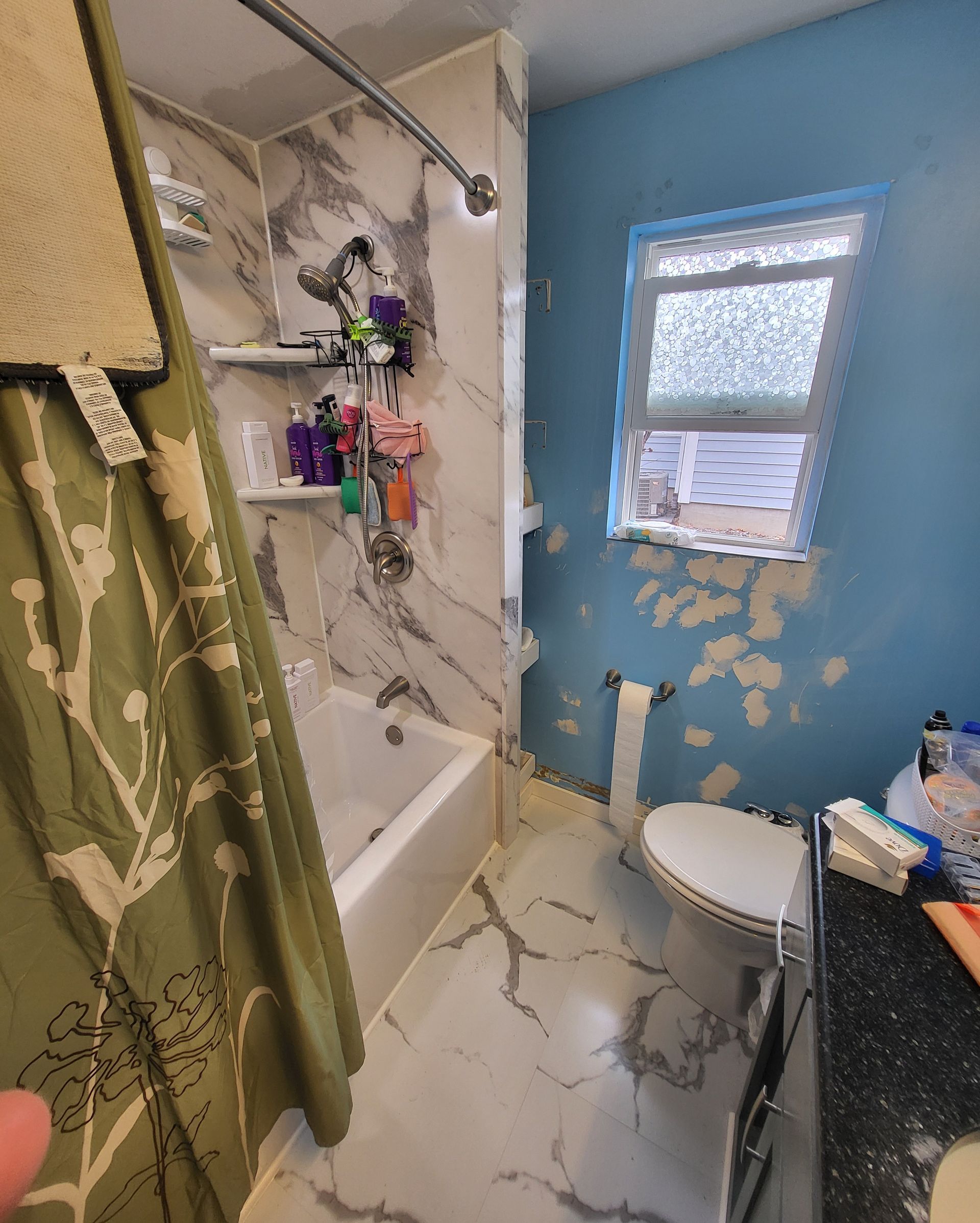 Bathroom with marble-look shower, blue walls, toilet, window with peeling paint, and green shower curtain.