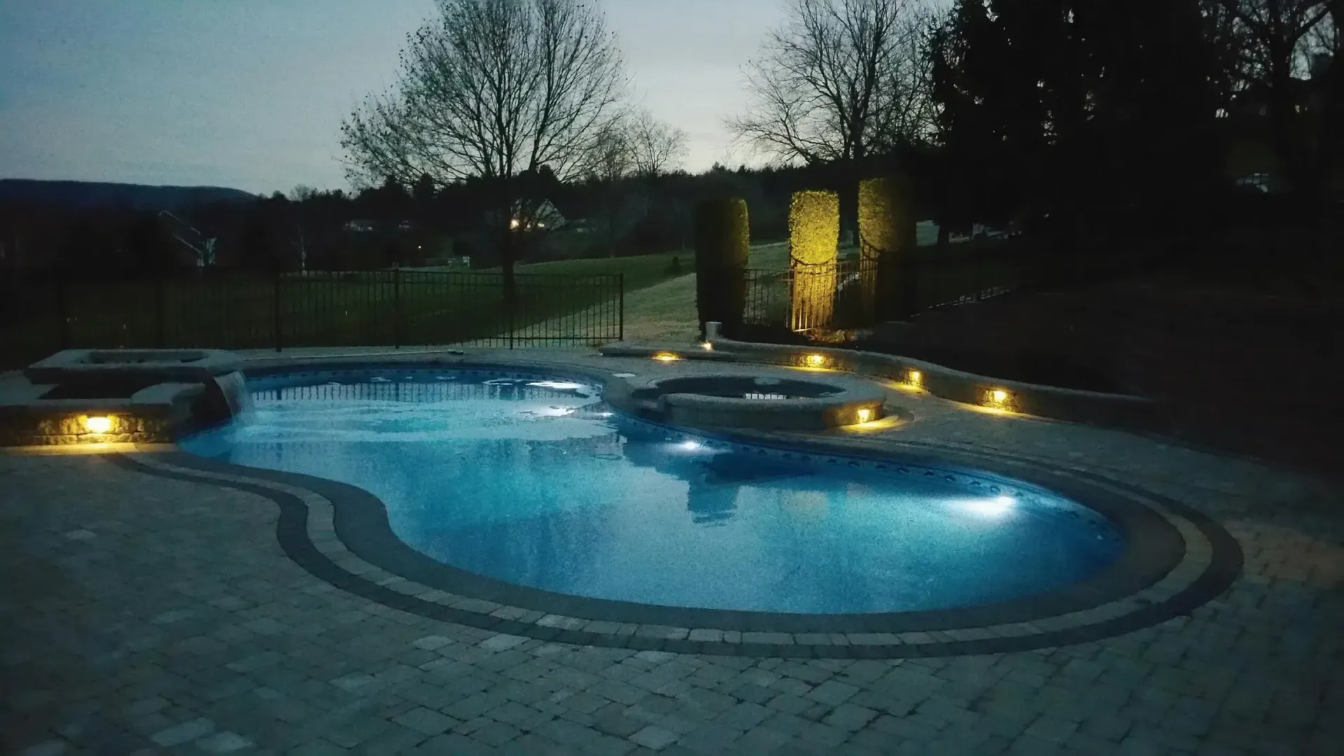 Illuminated pool at dusk with surrounding lights, brick patio, and landscaping.