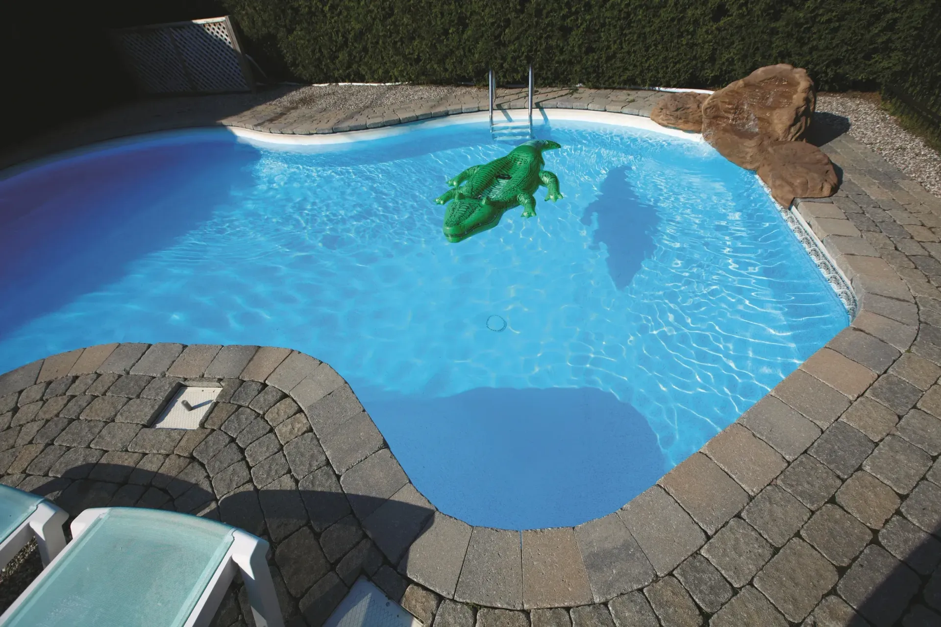 Swimming pool with green inflatable alligator. Stone border surrounds the blue water.