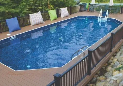 Oval in-ground pool surrounded by a wooden deck and fence. Pillows in blue, green, and striped patterns rest on the deck.