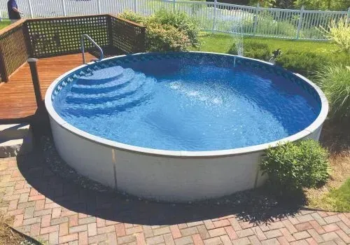 Round above-ground pool with steps, surrounded by brick patio, deck, and greenery.