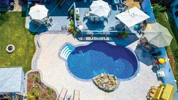 Aerial view of a backyard with a pool, deck, and umbrellas. Blue pool surrounded by stone patio.