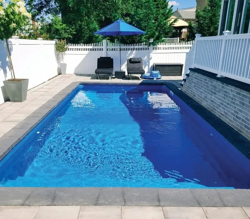 A rectangular blue pool in a backyard, with lounge chairs and an umbrella.