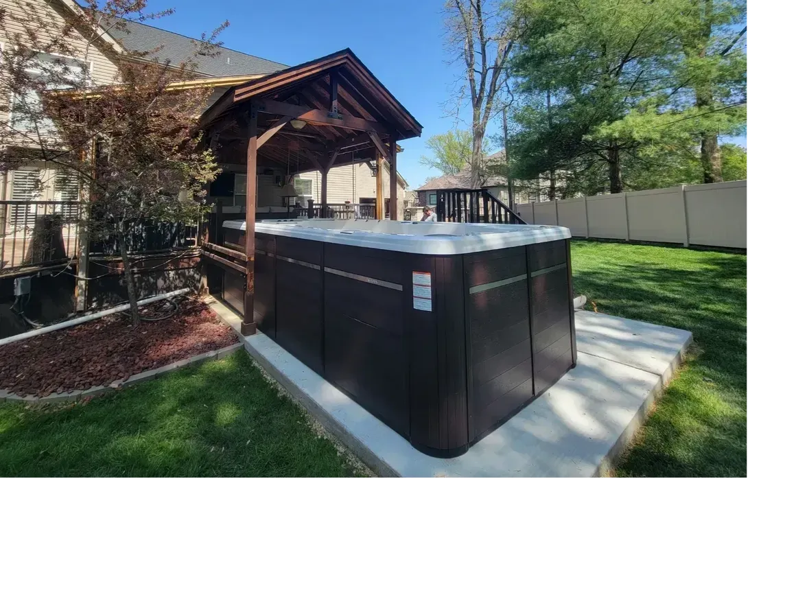 A dark brown hot tub with a gazebo is set on a concrete pad, in a grassy backyard, next to a house.