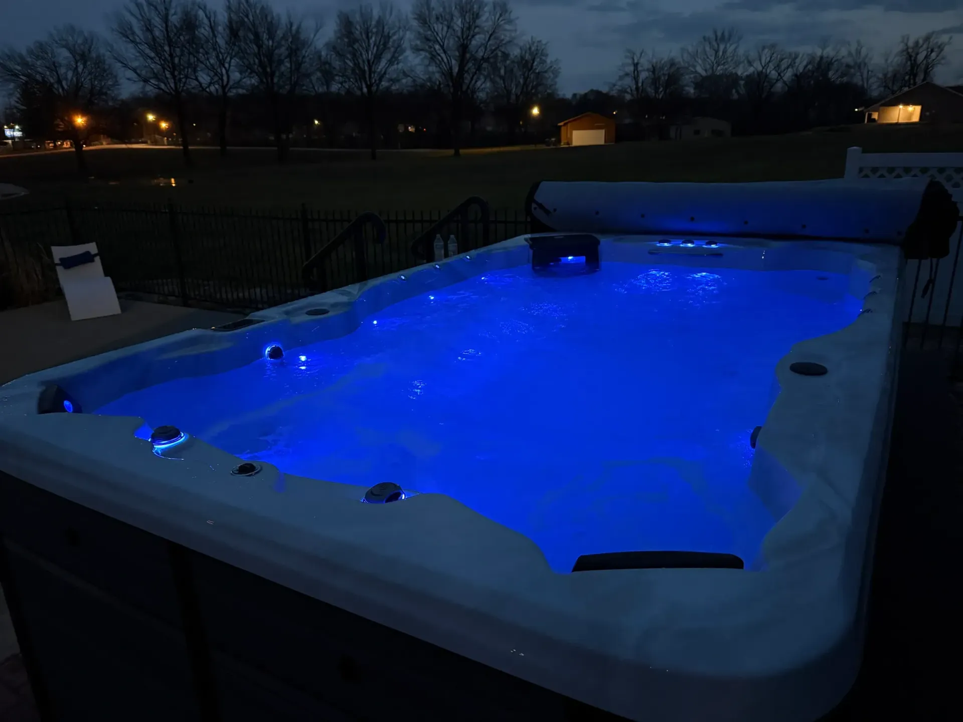 Lit blue hot tub at dusk in a yard, cover rolled back.