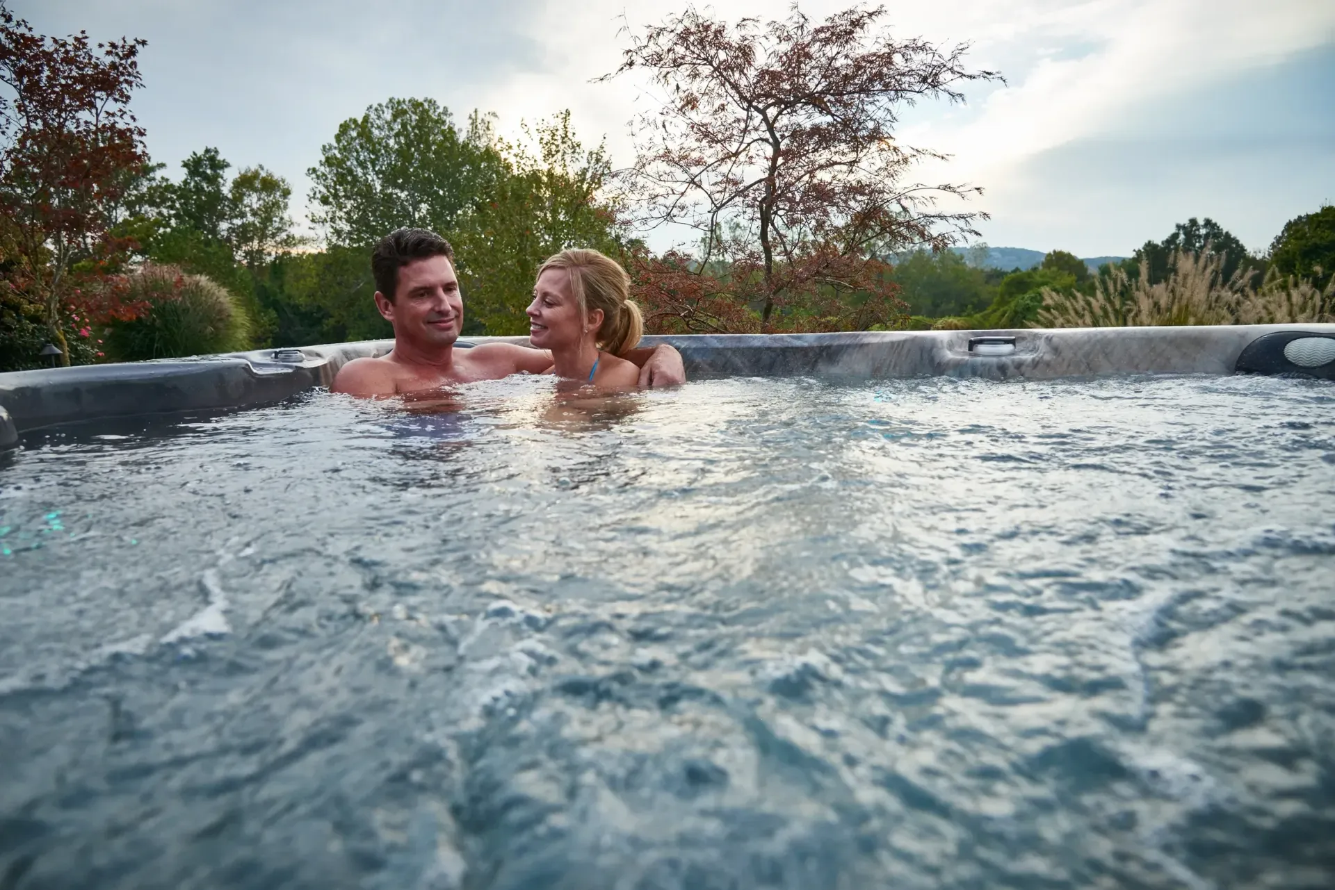 Couple in hot tub, looking at each other, with trees in the background.
