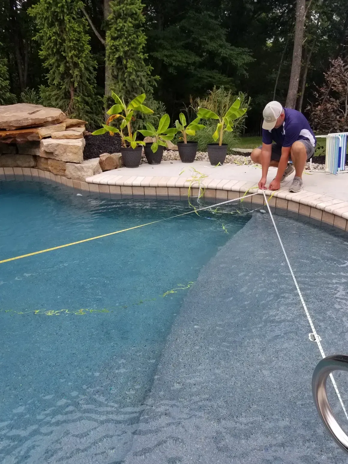 Person near pool edge adjusting yellow rope in pool. Plants in black pots on deck.