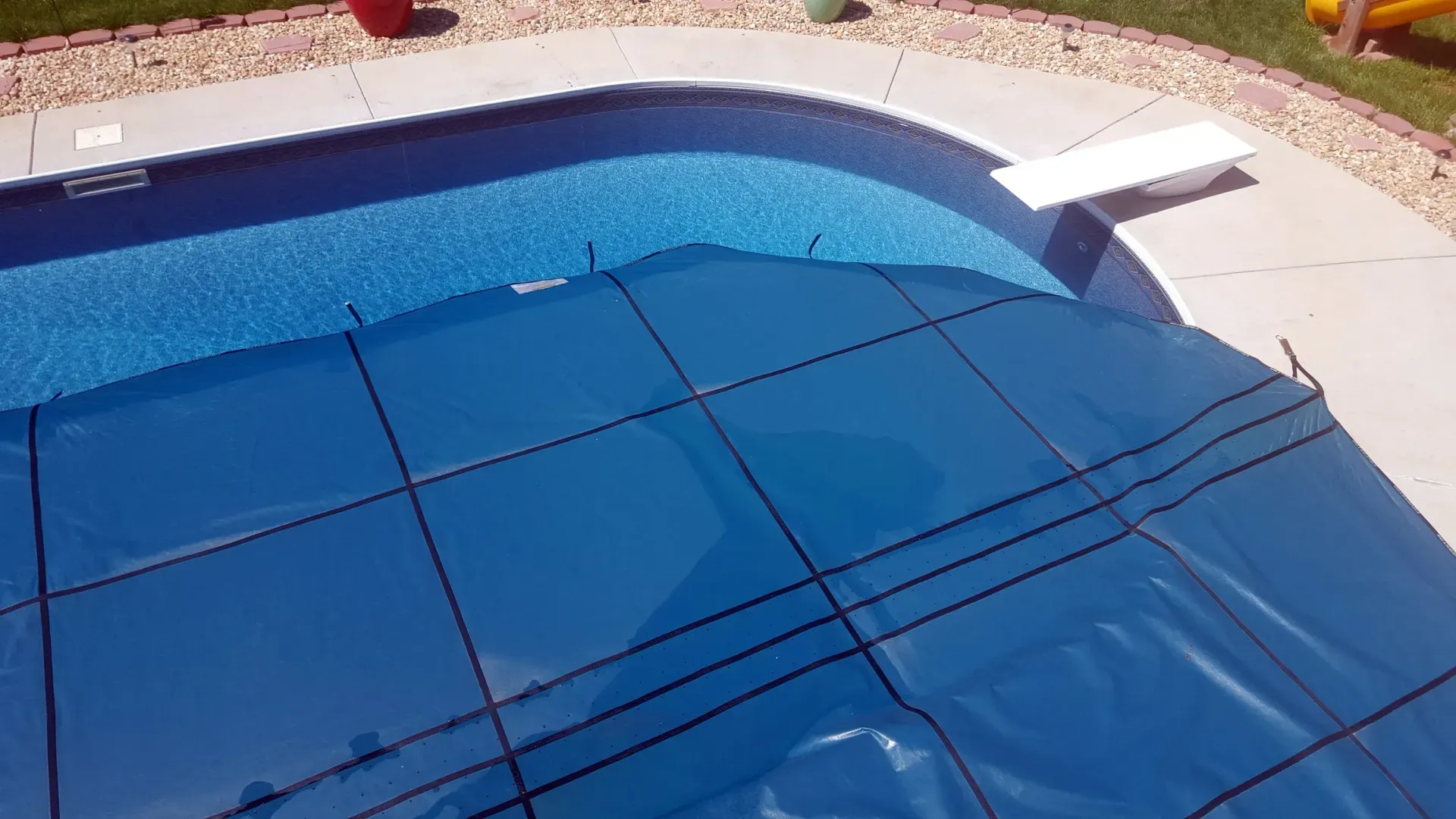 Blue pool covered with a blue mesh safety cover. Concrete deck, diving board, and landscaping visible.