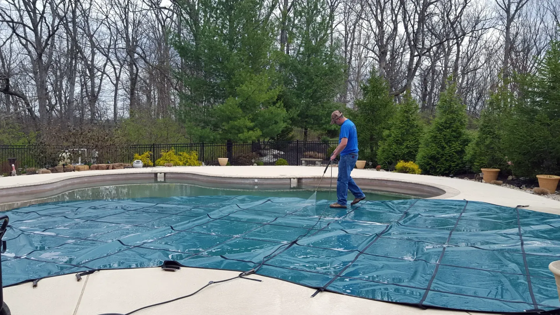 Man cleaning a blue pool cover with a hose, in a backyard setting. Trees in the background.