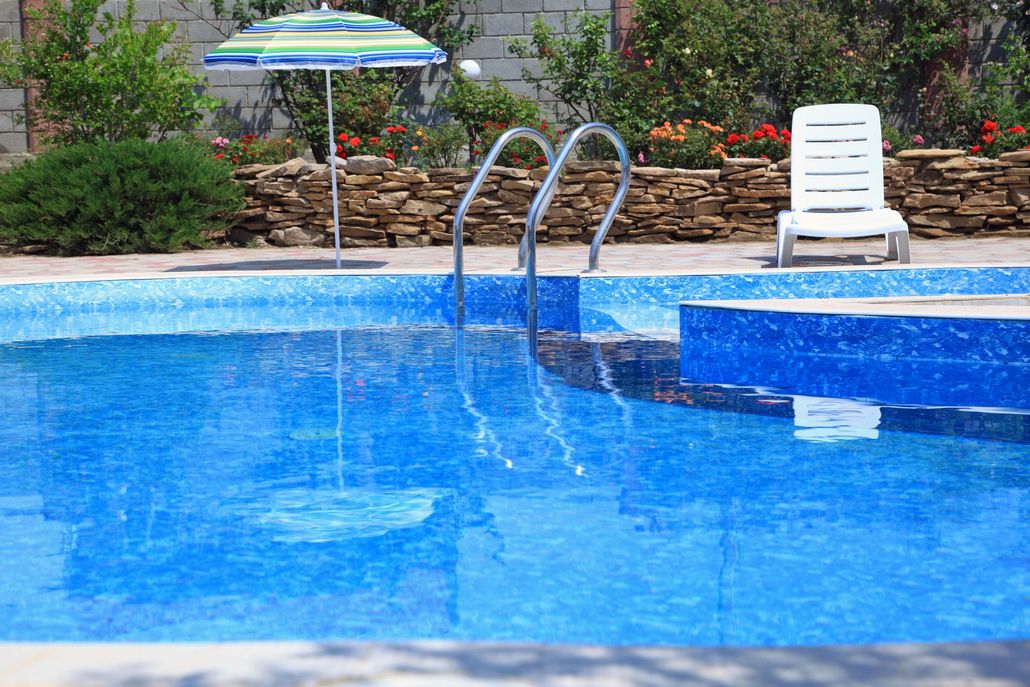 Blue-tiled swimming pool with ladder, umbrella, and lounge chair. Stone wall and bushes in background.