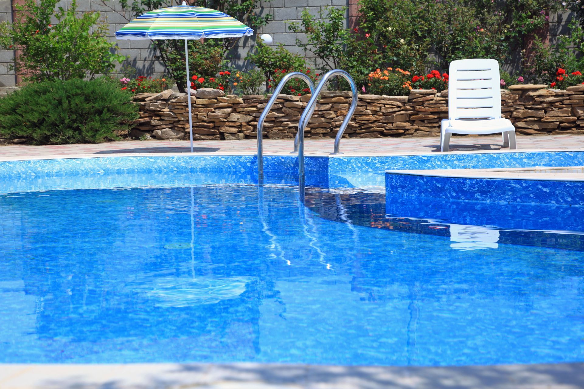 Blue-tiled swimming pool with ladder, umbrella, and lounge chair. Stone wall and bushes in background.