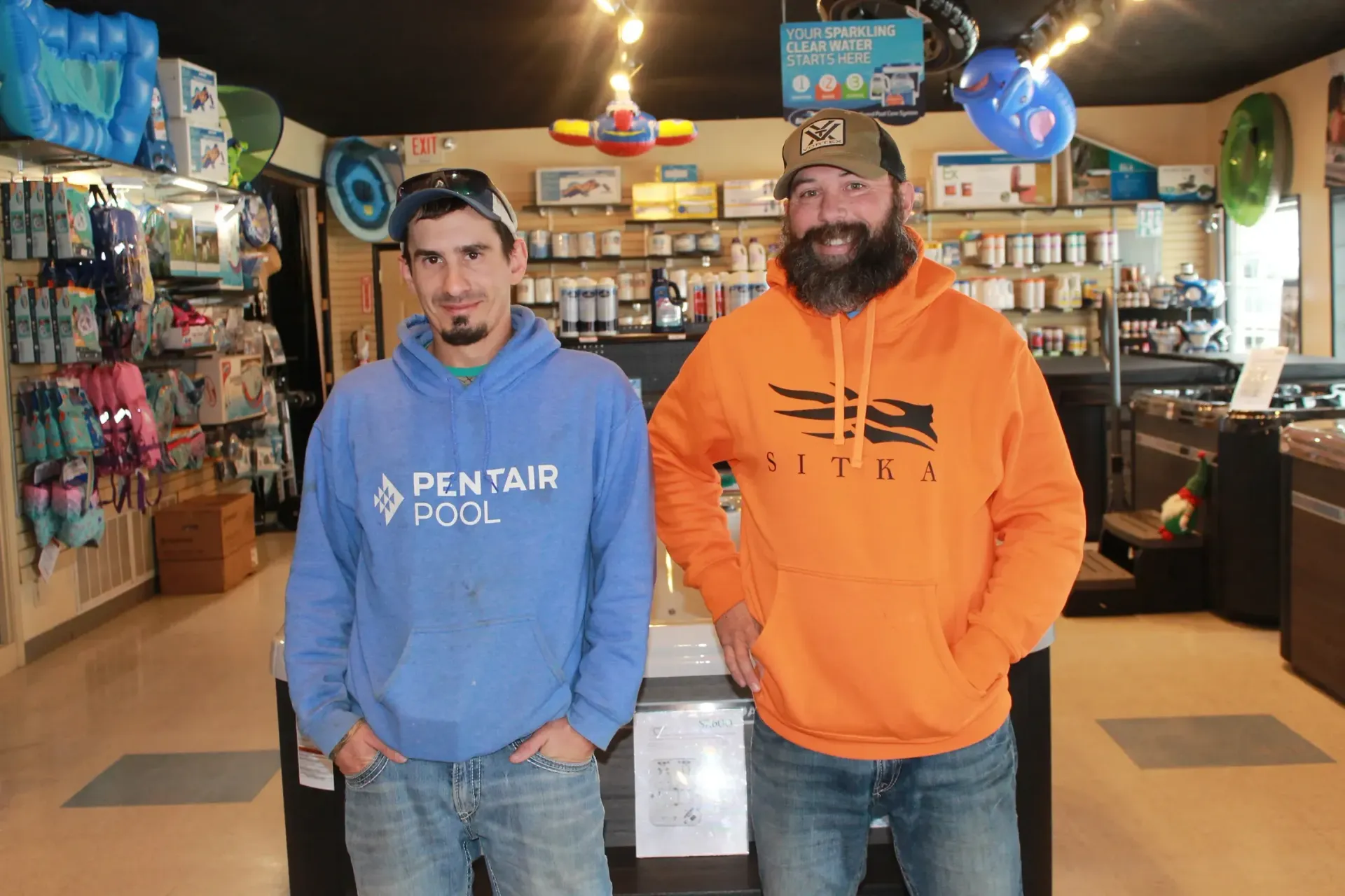 Two men in a pool supply store. One in a blue Pentair hoodie, the other in an orange sweatshirt, standing behind a counter.