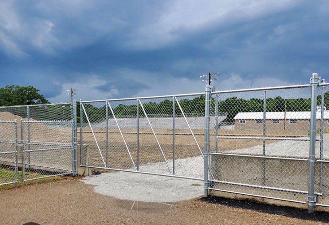 Chain link gate open, leading to gravel area with buildings and trees under a cloudy sky.