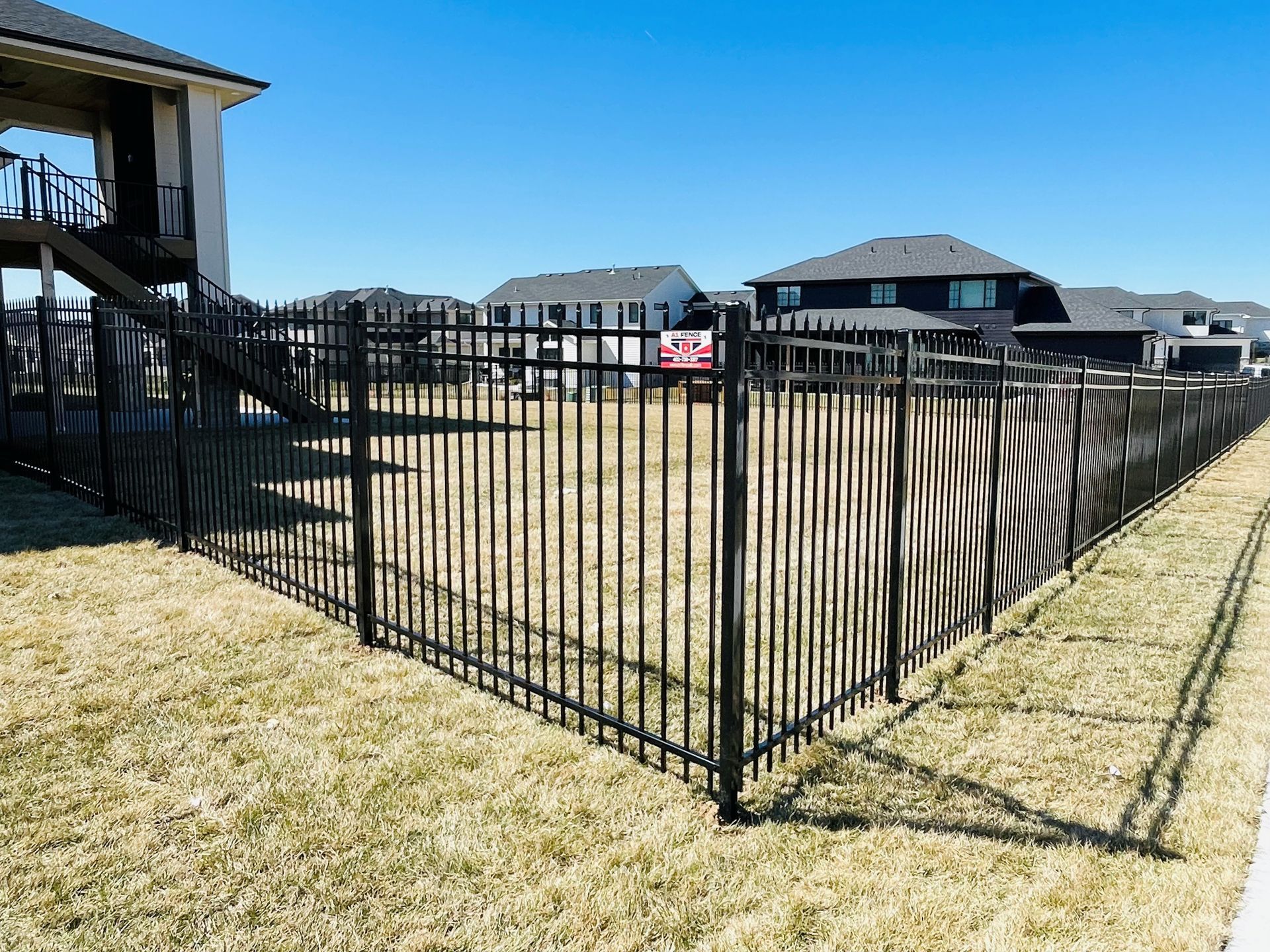 Black metal fence surrounds a grassy yard in a suburban neighborhood, blue sky.