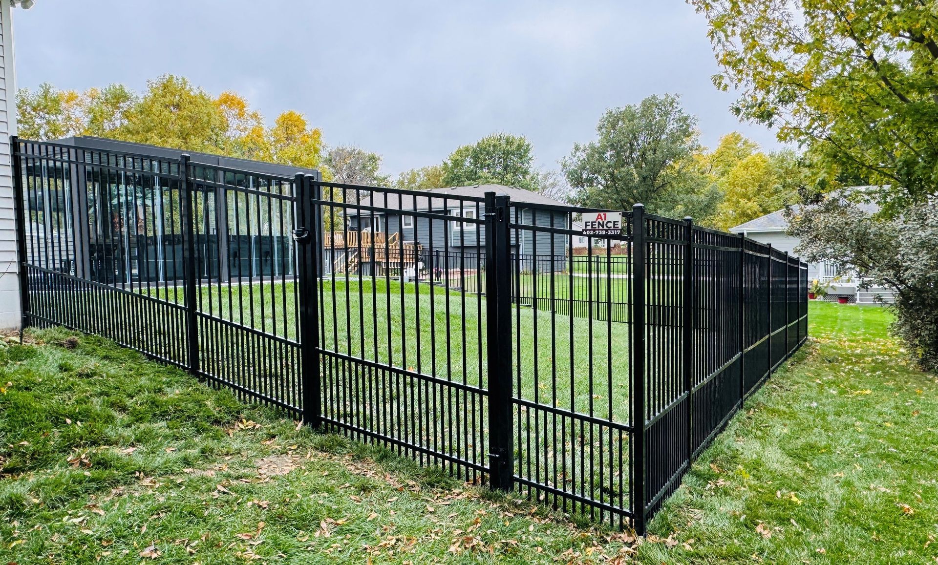 Black metal fence encloses a grassy backyard on a cloudy day, surrounding a home.