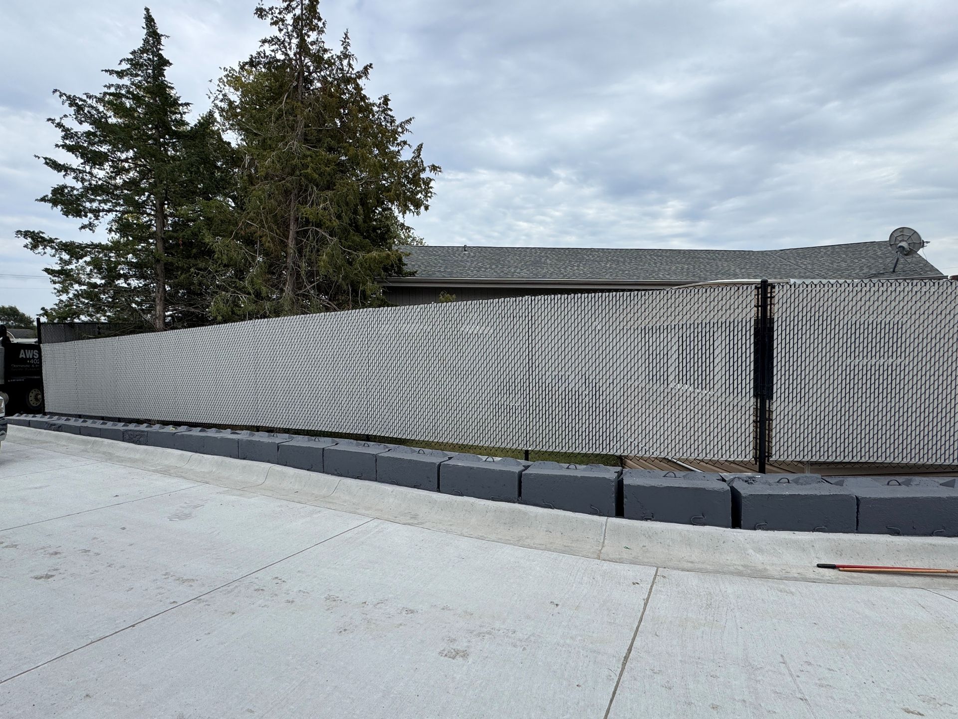 Gray patterned fence with dark gray concrete blocks along a paved area, with trees and a building in the background.