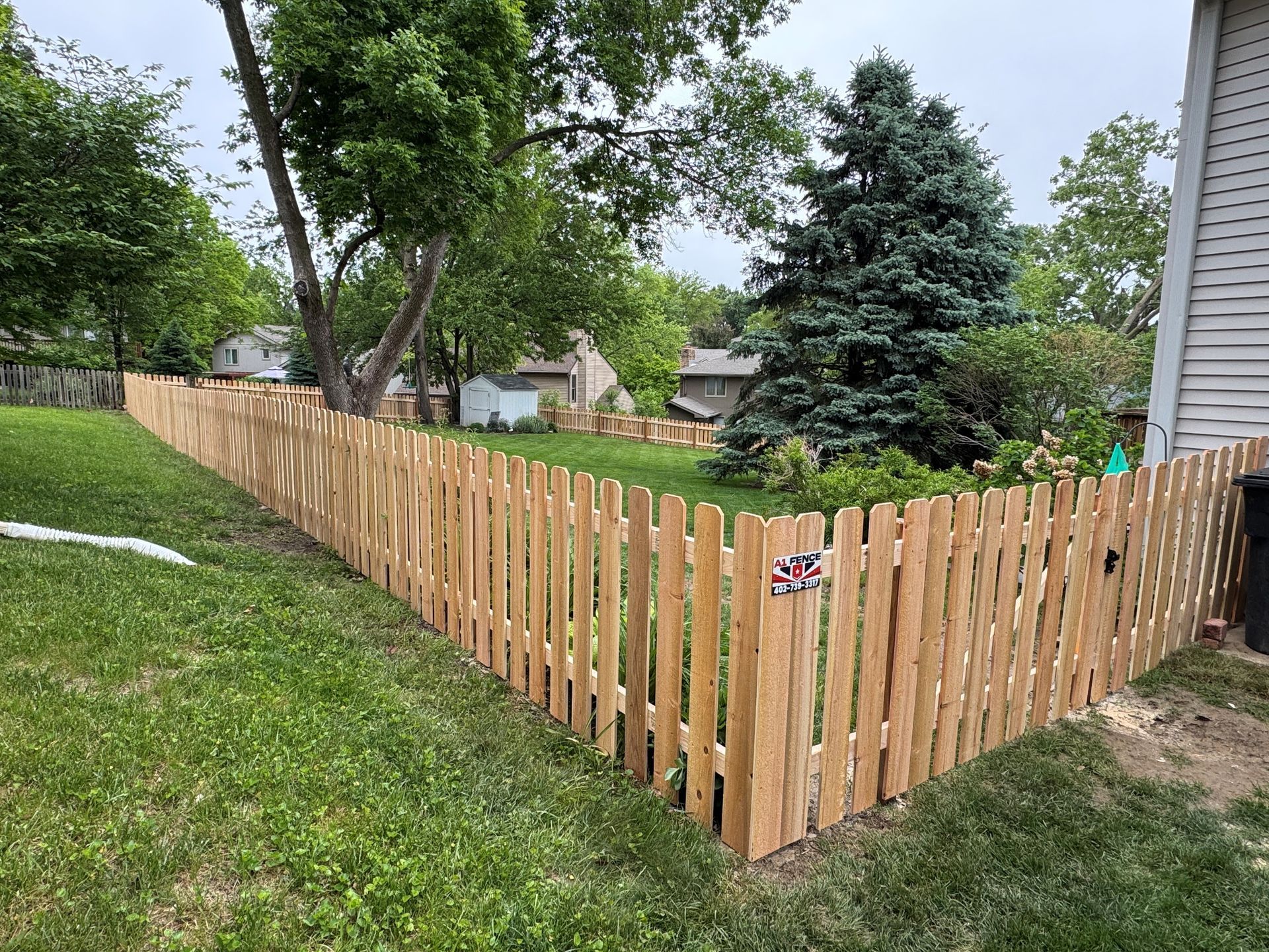 Wooden picket fence curves around a grassy yard, partially encircling a house and trees.