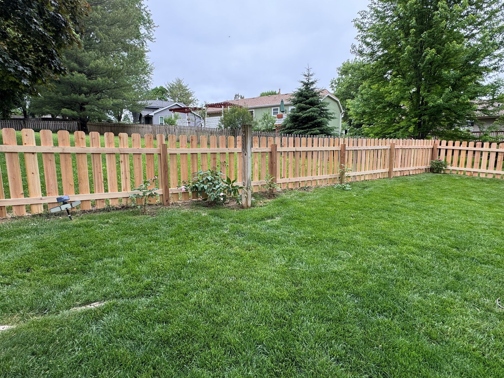 Wooden picket fence in a green backyard, with trees and a cloudy sky.