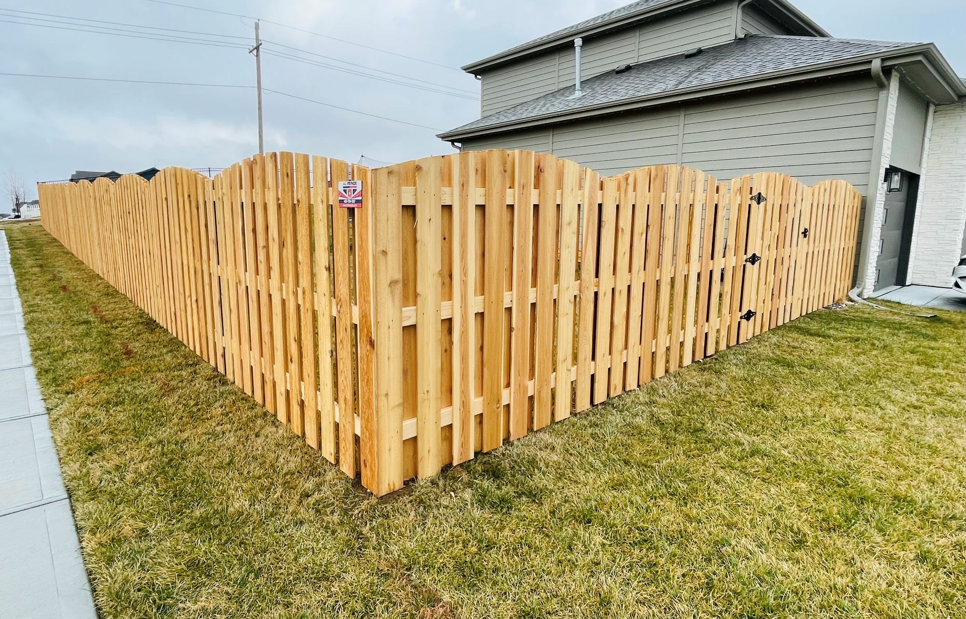 Wooden fence surrounding a grassy yard next to a house with a gray roof and a cloudy sky.