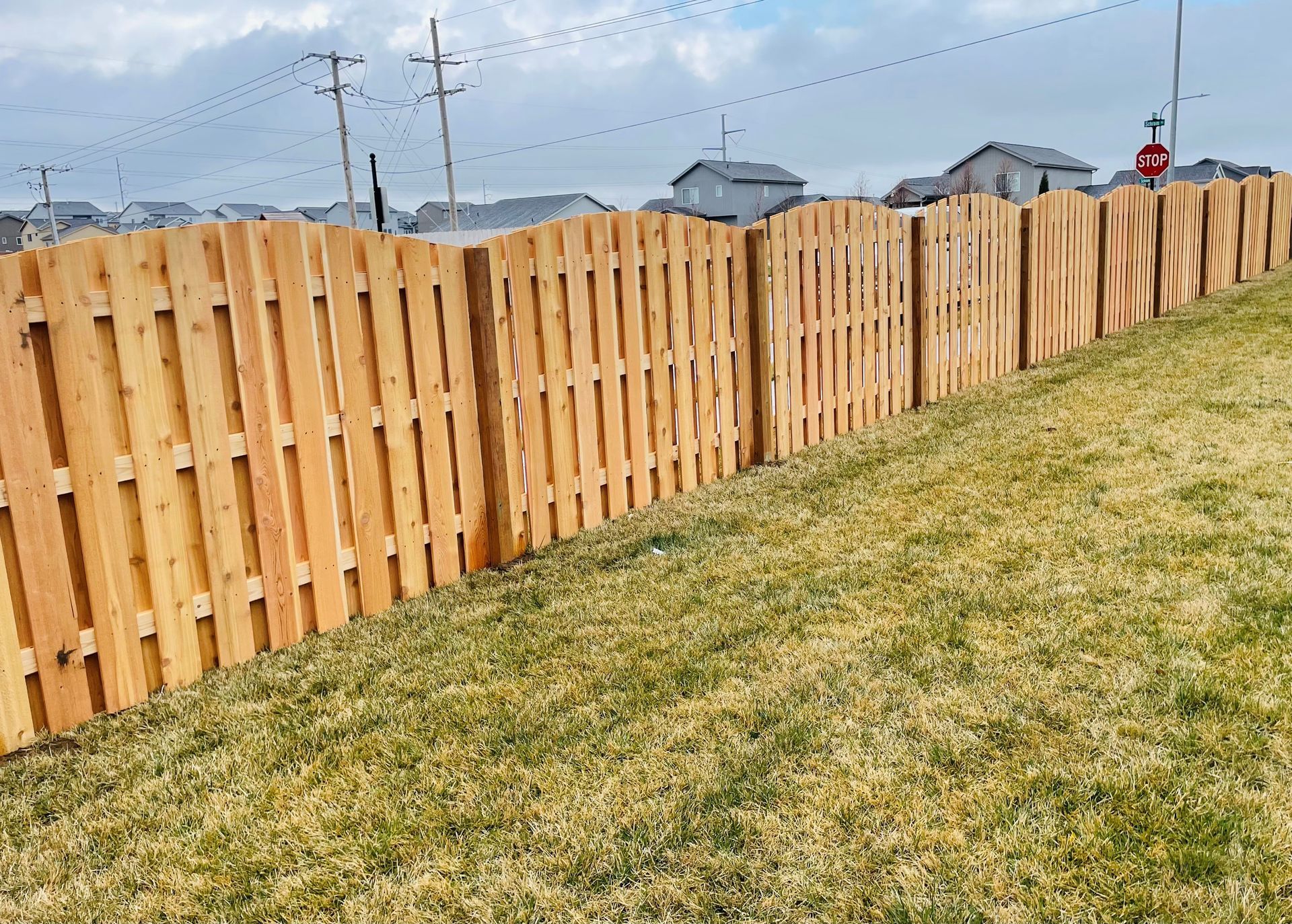 Wooden fence on a grassy hill, leading toward houses under a cloudy sky.
