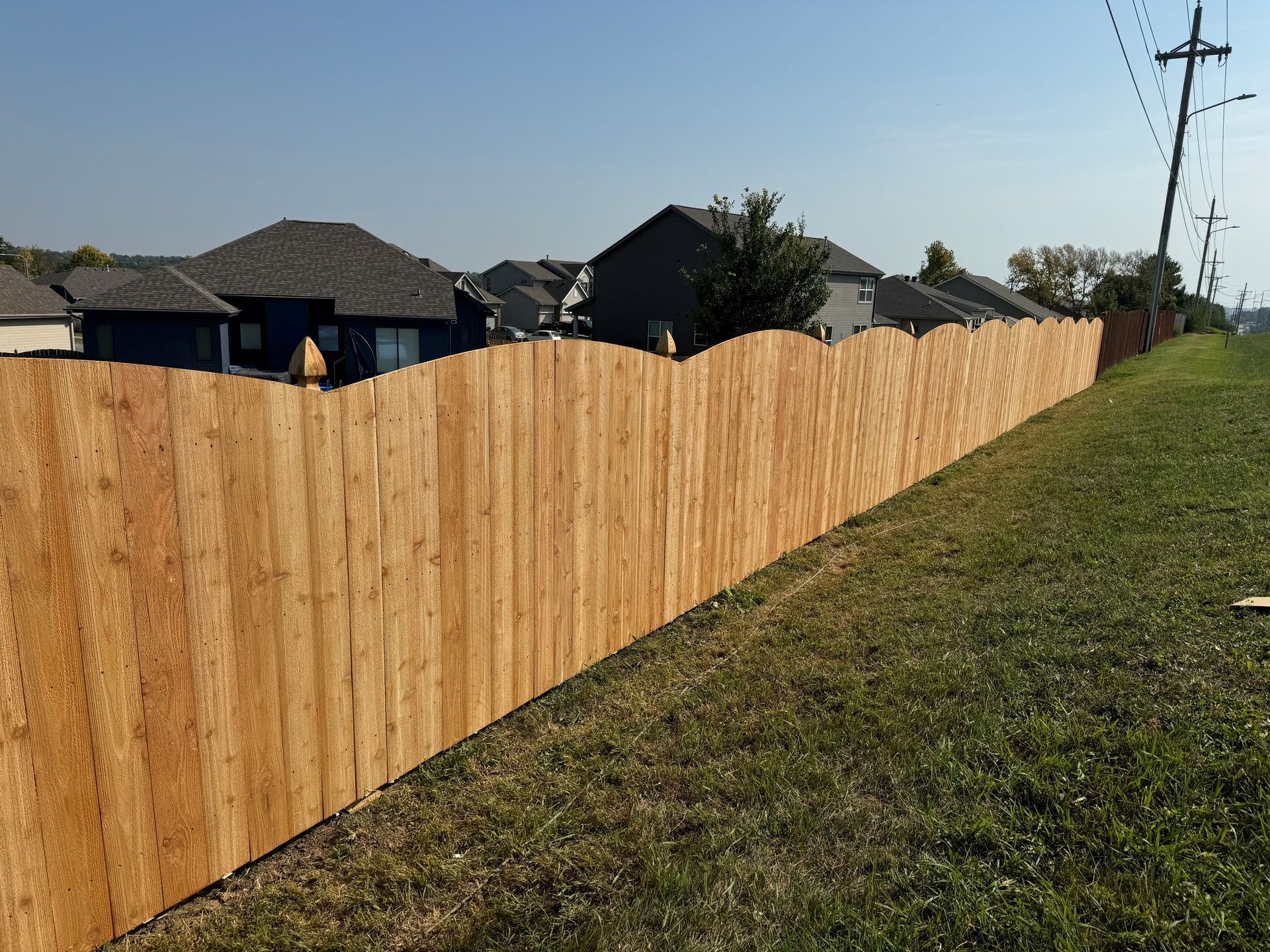 Wooden fence with a scalloped top, separating a grassy area from a suburban neighborhood under a blue sky.