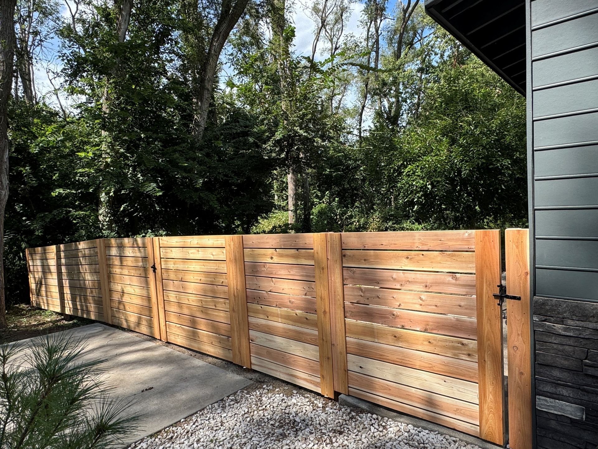 Wooden fence next to a house with a gravel path, surrounded by trees.