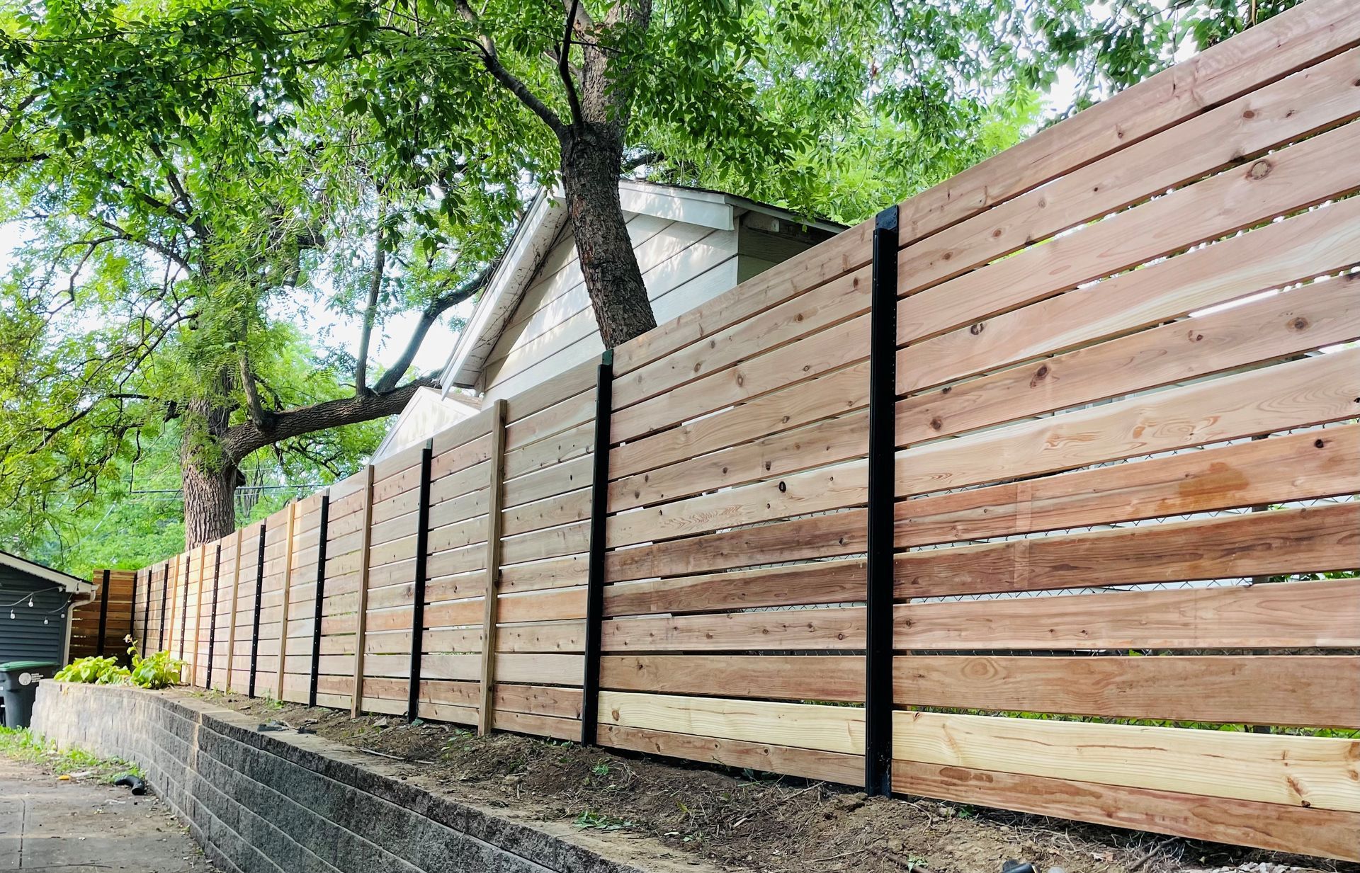 Wooden horizontal slat fence in front of a house, with black posts. Green trees in background.