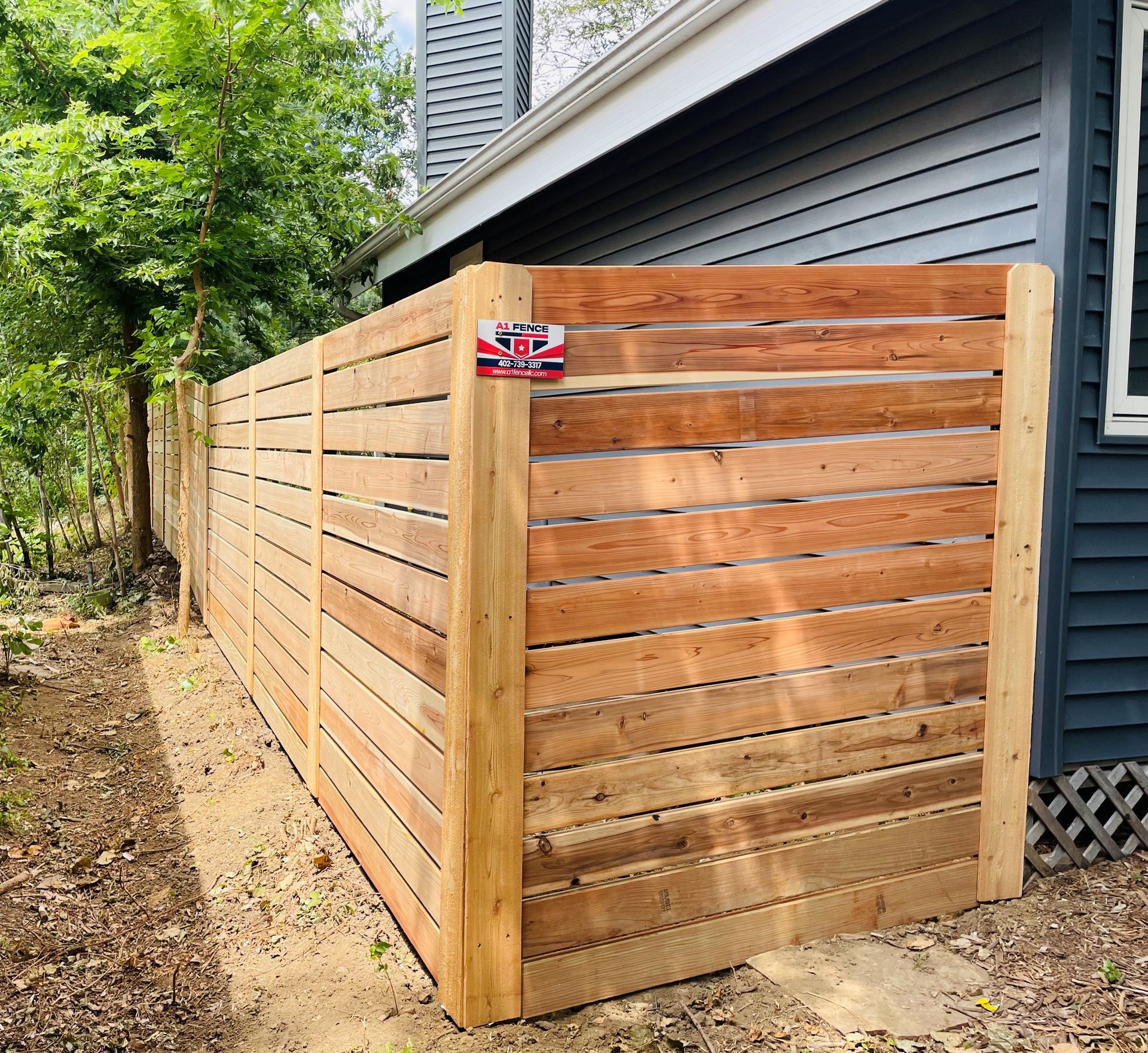 Wooden horizontal slat fence alongside a blue house, extending to the left in a yard.