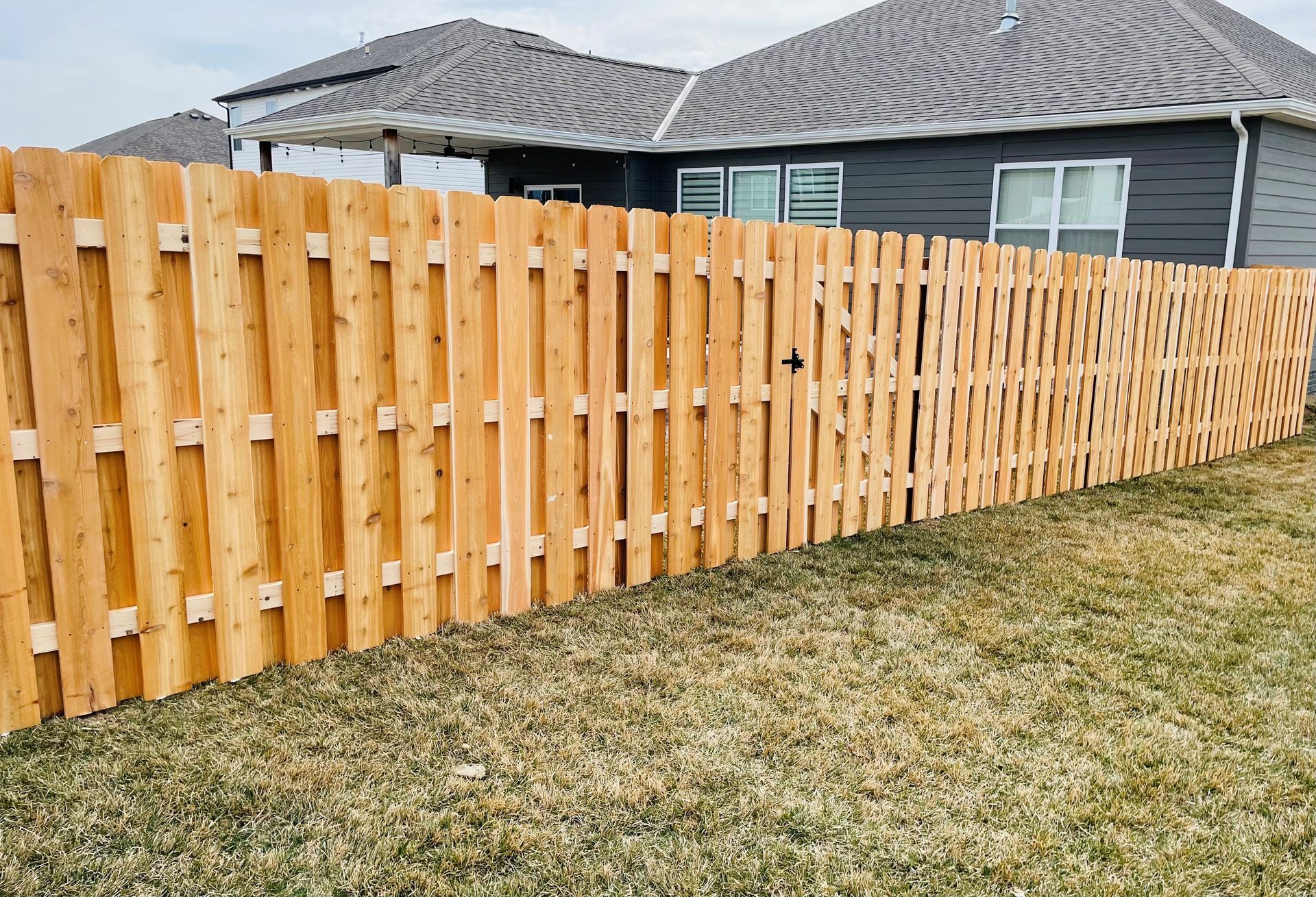 Wooden fence surrounding a house with gray siding and a gray roof the fence is on a grassy lawn.