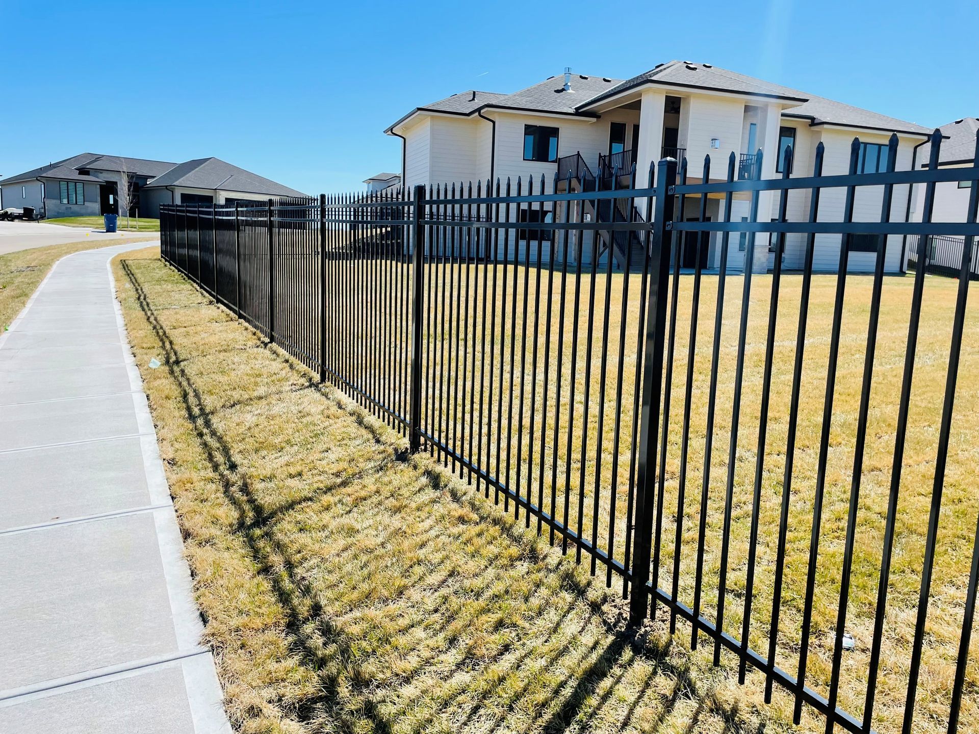 Black metal fence along a sidewalk, separating grass from houses on a sunny day.